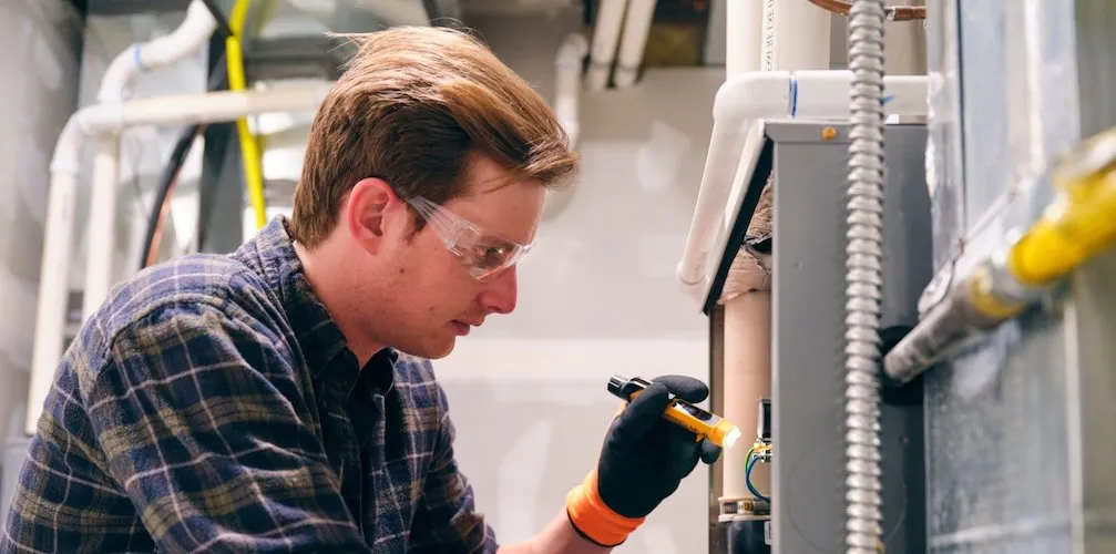 A technician wearing safety glasses and gloves inspecting the furnace. He is using a small flashlight to examine the internal components and electrical connections.