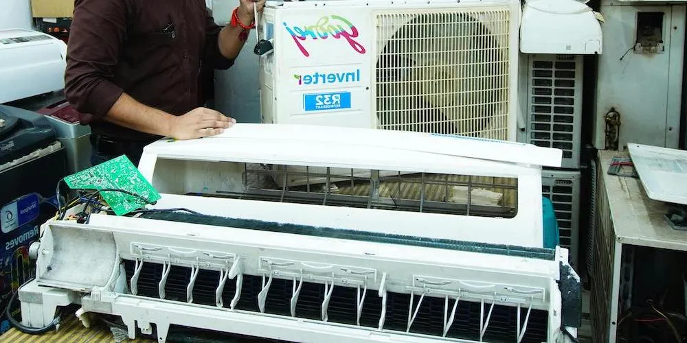 A technician in a workshop setting working on a disassembled air conditioner. The internal components, including the circuit board and the blower wheel, are exposed for deep cleaning or repair.