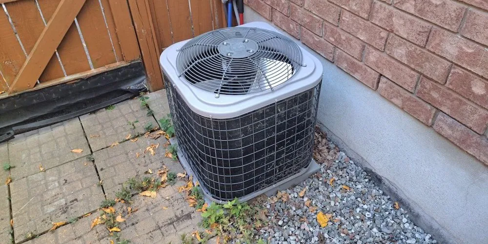 A square, grey outdoor central air conditioning unit sitting on a gravel surface next to a brick house. A wooden gate and stone pavers are visible in the background.