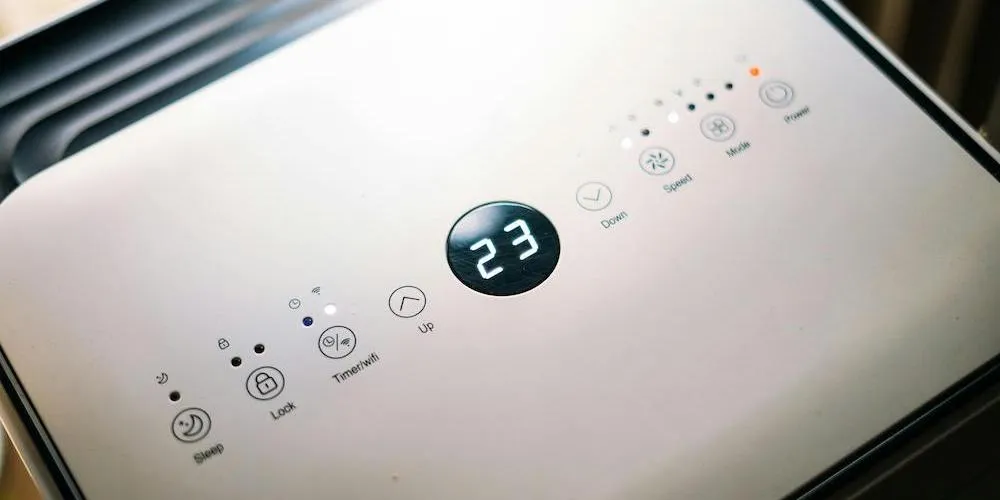 A close-up, top-down view of the white touch-control panel of a portable air conditioner or dehumidifier. The digital display in the center shows "23" in bright white LED numbers. Surrounding the display are touch-sensitive icons for Power, Mode, Fan Speed, Timer/Wi-Fi, Lock, and Sleep mode.